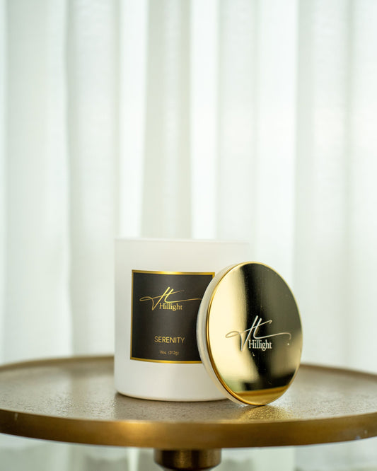 Candle with gold lid and label on a round wooden surface with white curtains in the background