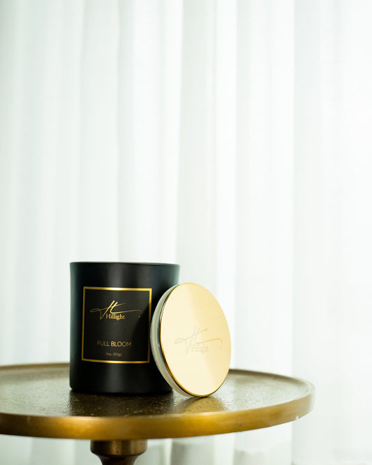 Black candle with gold lid on a wooden table against a white curtain background