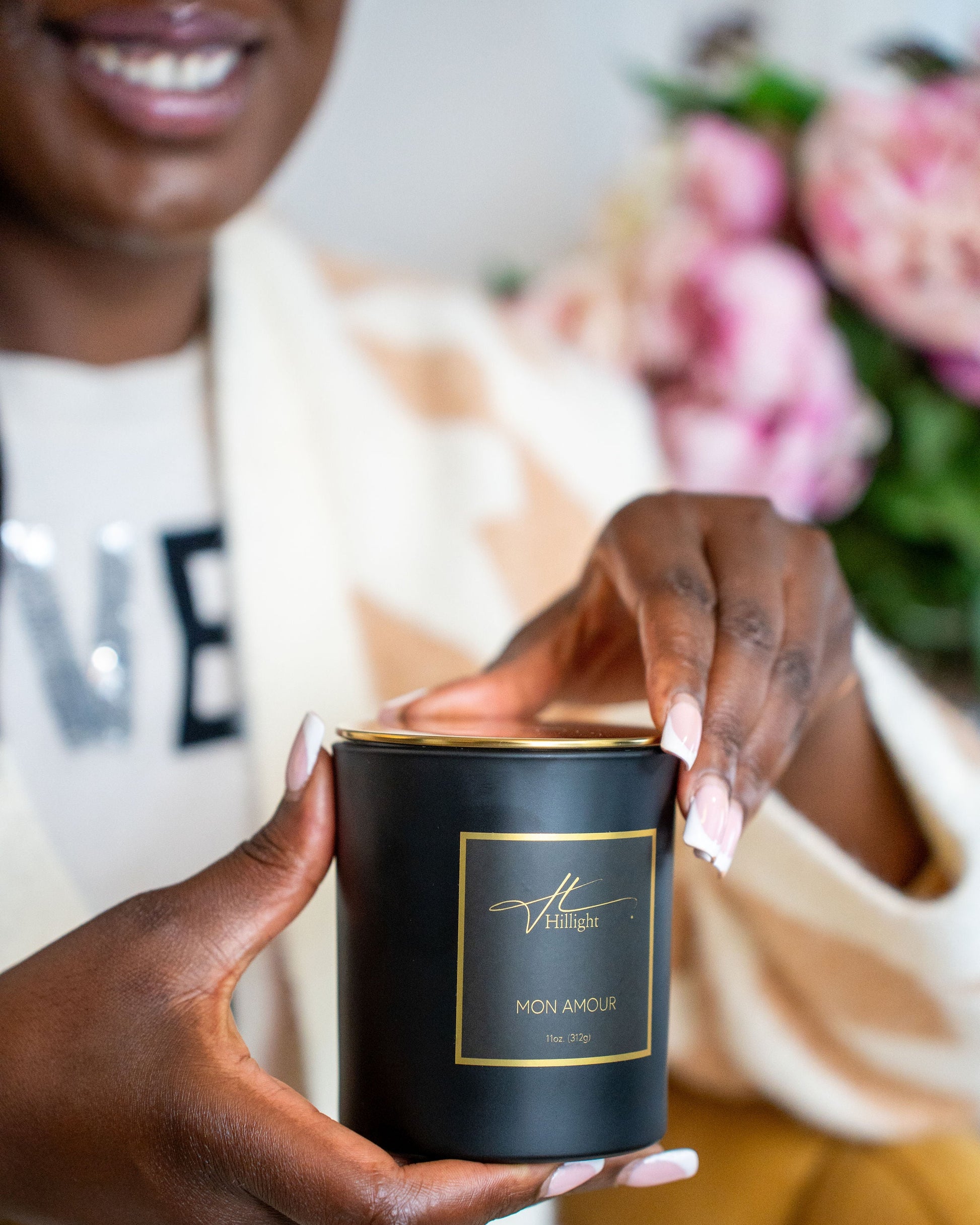 Person holding a black candle labeled 'Mon Amour' with flowers in the background